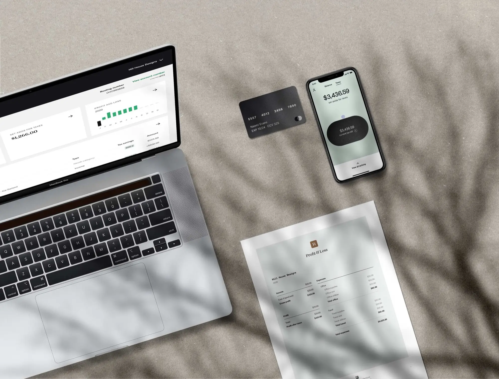 A laptop showing the Found dashboard beside a black Found debit card, a phone showing $3,436.59 set aside for taxes and a printed profit and loss statement, on a sand-coloured surface under tree shadows