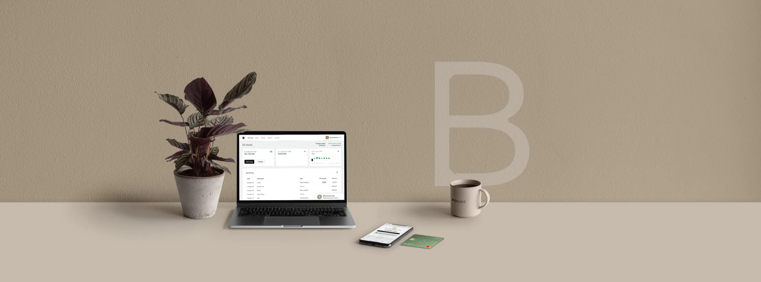 A laptop showing the Found app beside a potted calathea, a phone, a green Found debit card and a mug, on a taupe desk against a wall carrying a large letter B