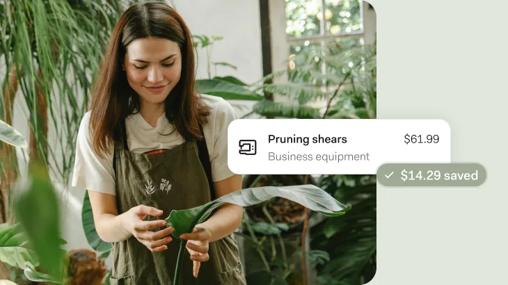 A woman in a white t-shirt and olive green apron with botanical embroidery tends to plants in a bright, plant-filled indoor space. She's holding a large green leaf while surrounded by various houseplants near a window. An overlay shows pruning shears listed as business equipment for $61.99 with $14.29 saved.