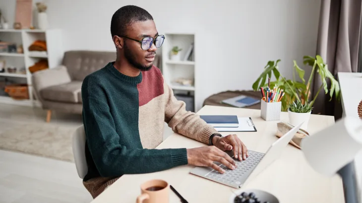 A man in a colour-block jumper typing on a laptop at a desk with a clipboard, potted plants, and a mug of coffee