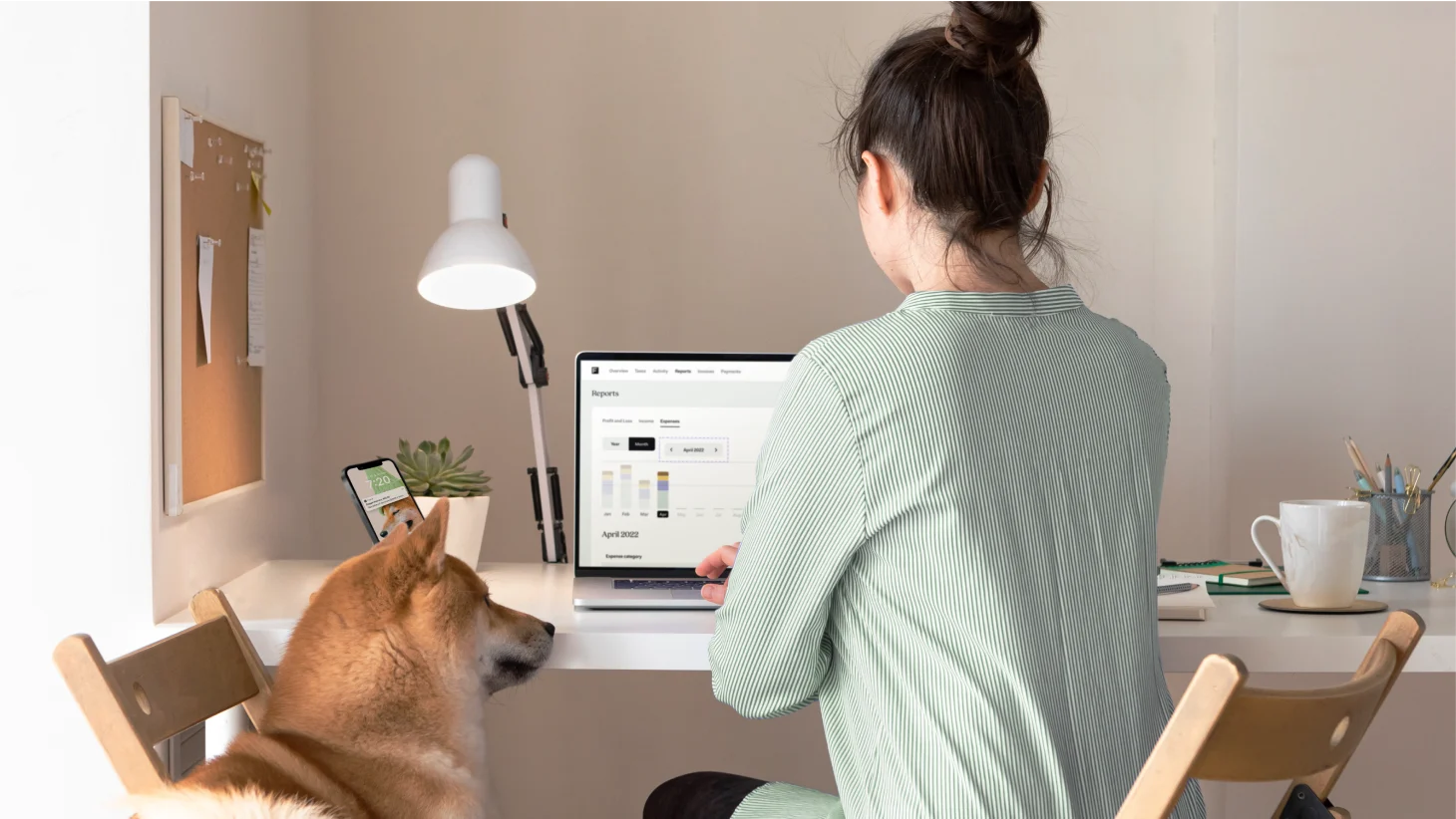 A woman working at a laptop showing Found's Reports screen with an April 2022 expense chart, with a phone and a dog nearby on a home-office desk