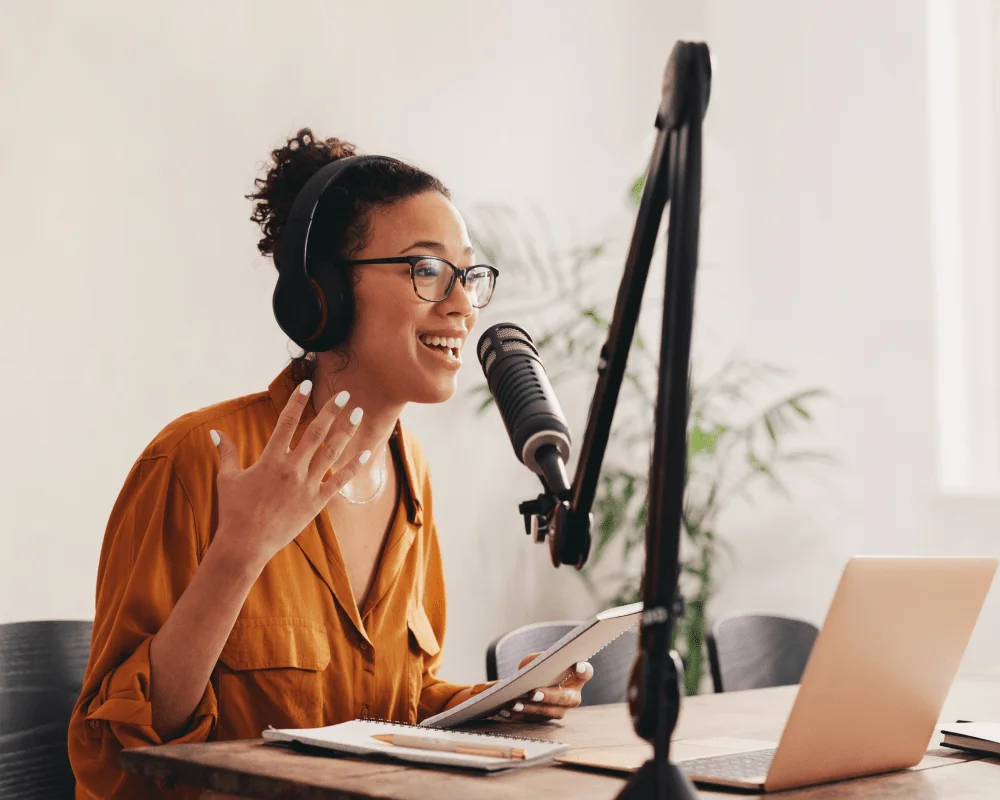 A podcast host in headphones talking into a studio microphone with notes and a laptop in front of her