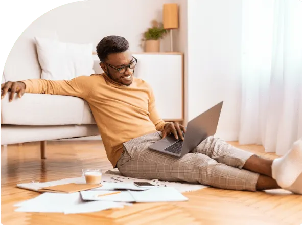 A man in an orange sweater sits on his living room floor working on a laptop, with paperwork and a coffee spread out beside him